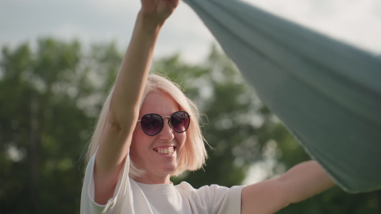 Playful mom wearing dark sunglasses swaying picnic blanket over heads with big smile, warm sunlight filtering through trees, casual white shirt, green park background, joyful family prep moment