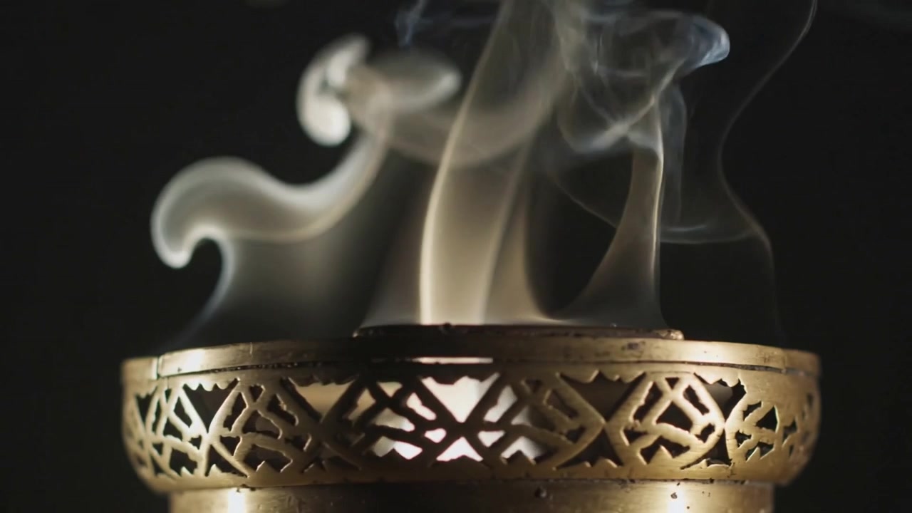 Emitting brass incense burner creating smoke from warm glow in studio, curls widening into ribbons