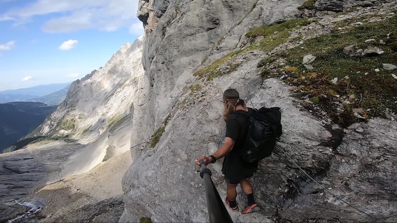un hombre joven, fuerte y en forma con cabello largo y tatuajes está agarrando una cuerda con una mano para guiarse por el camino muy estrecho y empinado en una gran roca