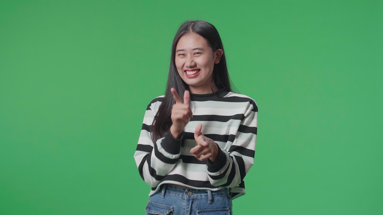 una mujer asiática sonriente mostrando un gesto a la cámara mientras está de pie frente al fondo de la pantalla verde