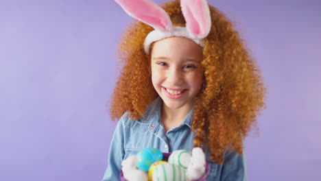 studio shot of girl wearing rabbit ears and holding basket of easter eggs against purple background