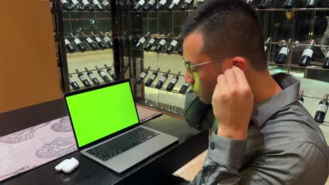 Wealthy-Asian-Guy-Wearing-Earbuds-and-Enjoying-Music-In-Front-of-Laptop-with-Green-Screen