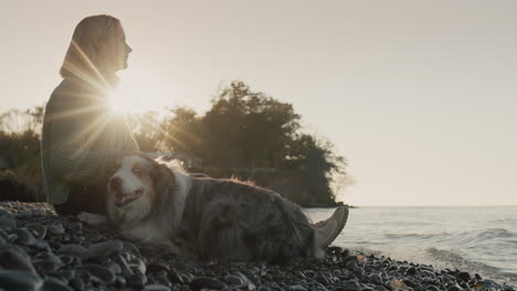 una mujer con un suéter cálido se sienta en la orilla de un lago al lado de un perro. el sol poniente lo ilumina y el surf