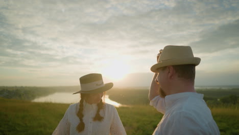 a couple stands in a grassy field at sunset as the man in a white shirt and hat playfully takes off his hat, bows, and then puts it back on while the woman in a white dress watches with a smile