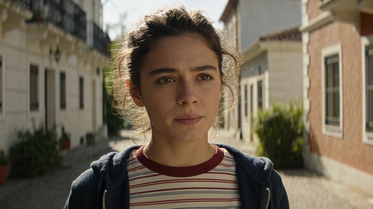 Donna con felpa con cappuccio che sposta lo sguardo, si ferma, scruta un vicolo in pietra con piante in cerca di direzione, mentre la telecamera si allarga.