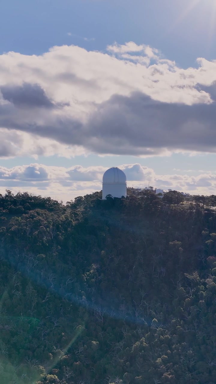 Droneoptagelse panorerer jævnt hen over en skovklædt bjergryg og afslører en stor observatoriekuppel under delvist skyet himmel med naturligt sollys og lens flare