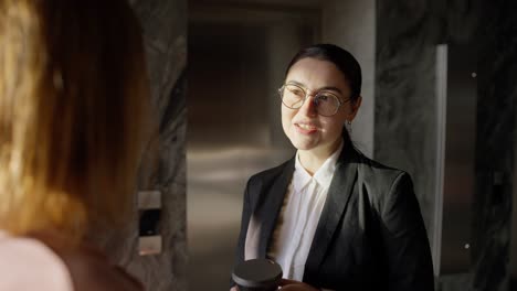 over the shoulder a confident brunette girl in round glasses and a black business uniform communicates with her colleague while drinking coffee in a modern sunny office during work