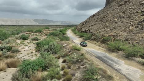 Aerial-drone-shot-flying-over-a-car-driving-along-a-road-at-the-foothill-of-a-mountain-range-in-Hingol,-Balochistan,-Pakistan-on-a-cloudy-day