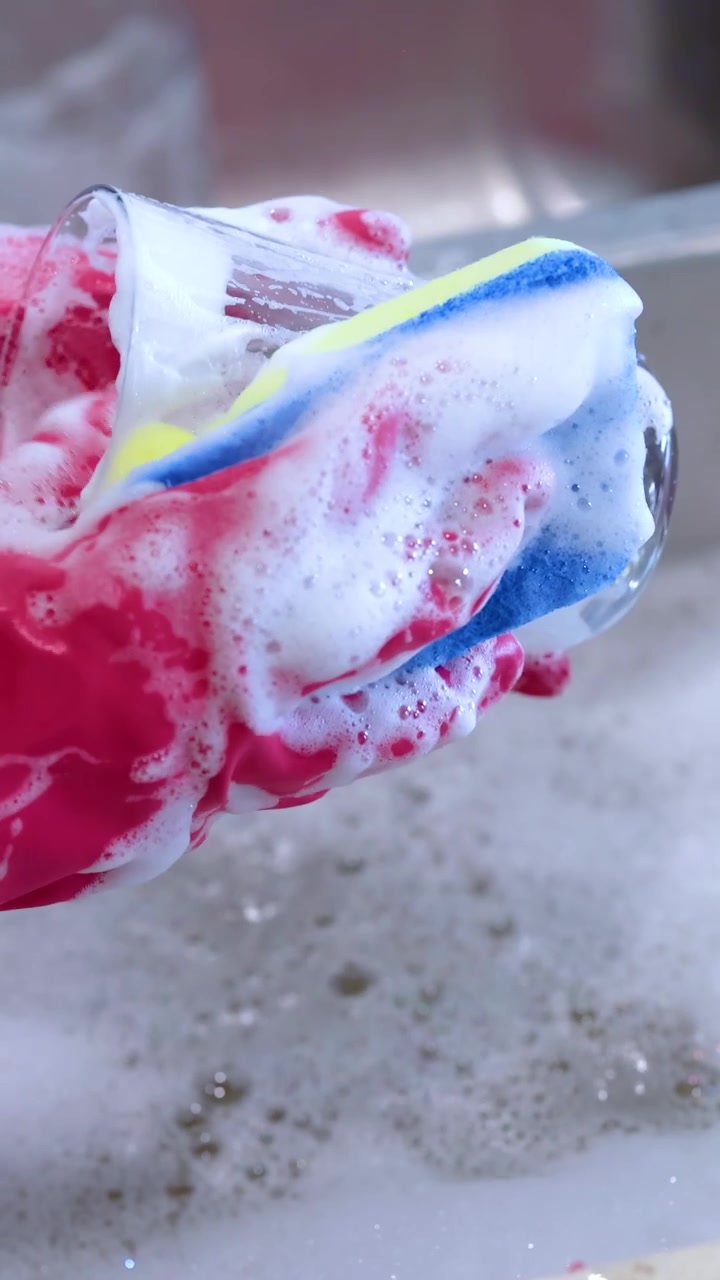 A person wearing red gloves cleans a glass with a sponge in a bubbly sink