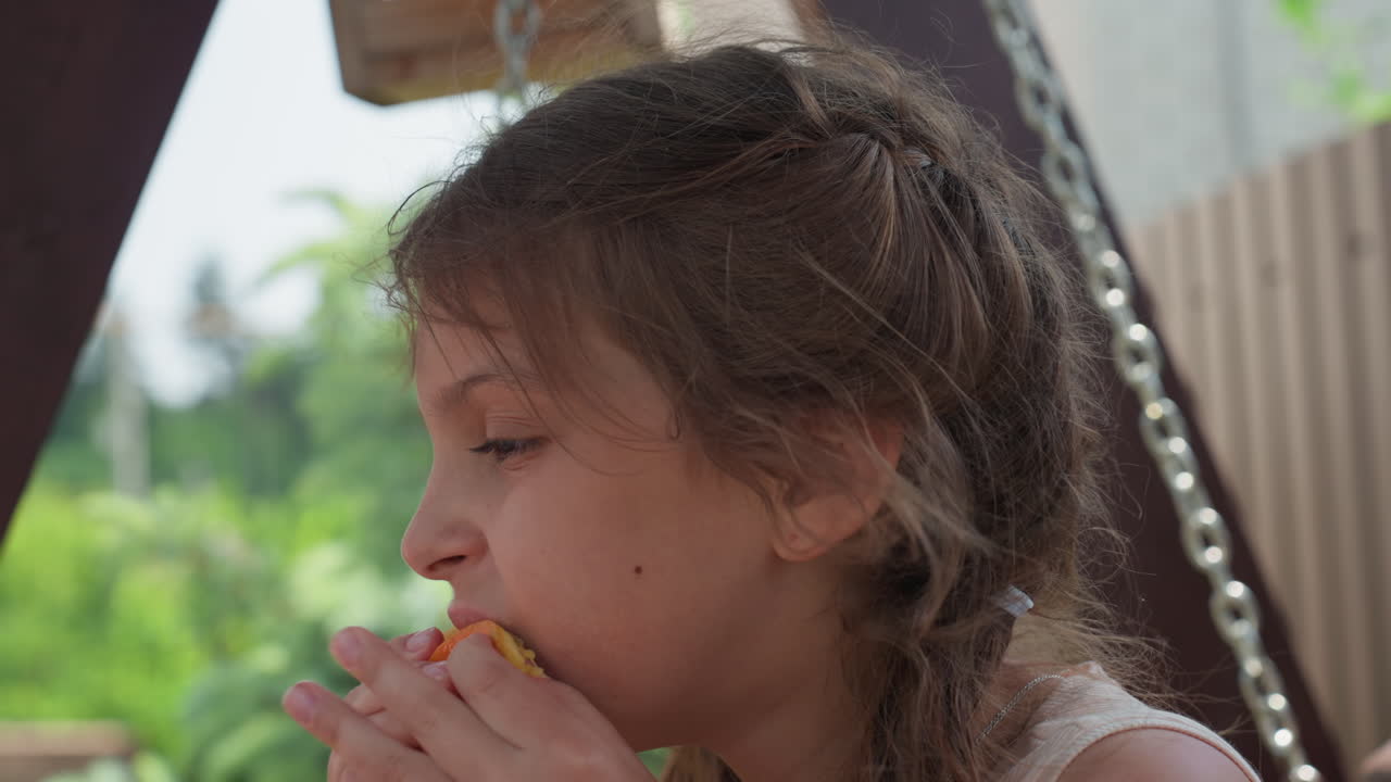 Girl On Swing Savoring Tangy Orange Drink, Child Delights In Biting Juicy Citrus Outdoors In Garden Swing, Young Girl Enjoys Drinking And Tasting Fresh Orange Juice While Sitting On Garden Swing