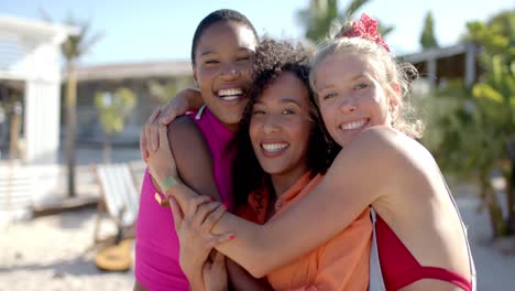 portrait of happy diverse female friends embracing on beach with beach house and palm trees