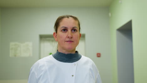woman in white coat walks with straight face through clean indoor walkway appearing focused and calm overhead lights reflect off glossy walls background shows emergency exit and green plant