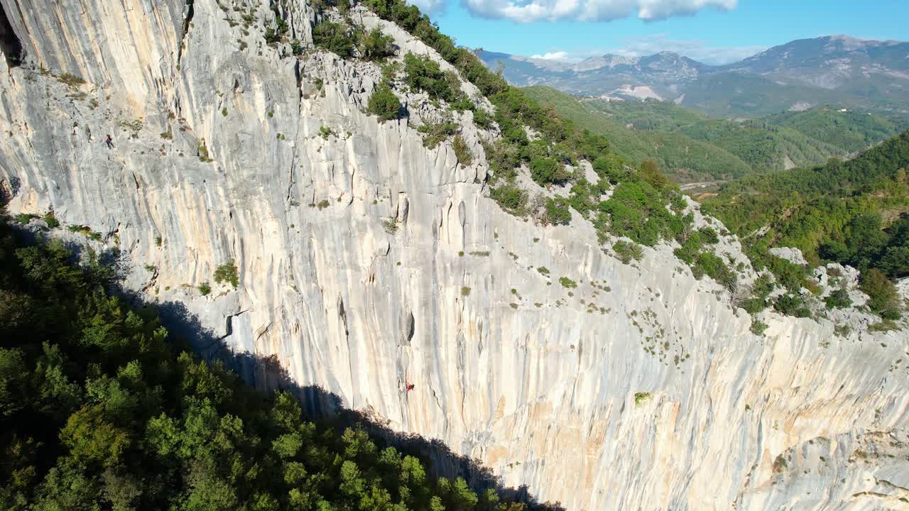 Aerial shot (pan, tracking) of a climber on the rock face in Brar, with the mountainous landscape in the background. Highlights training, technique, and the sport of climbing