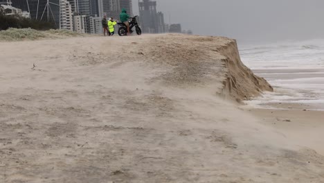 A close-up view of a sandy cliff edge with city buildings in the background, highlighting coastal erosion.