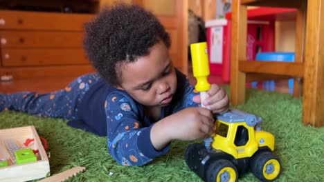 three-year-old back child fixing his toy bulldozer lying on a green rug at home, wearing a blue pjyamas