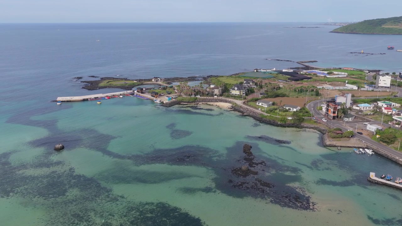 Horisontalt utsnitt som panorerer mot venstre over Sinheung-ri nær Hamdeok-stranden, Jeju-øya, Sør-Korea, som viser turkist vann, vulkanske steiner, kystlinje, landsby og jordbruksland