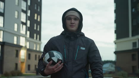 atleta confiado sosteniendo una pelota de fútbol y levantando los pulgares durante el entrenamiento en la arena urbana, mostrando determinación con edificios residenciales modernos en el fondo durante la noche nublada