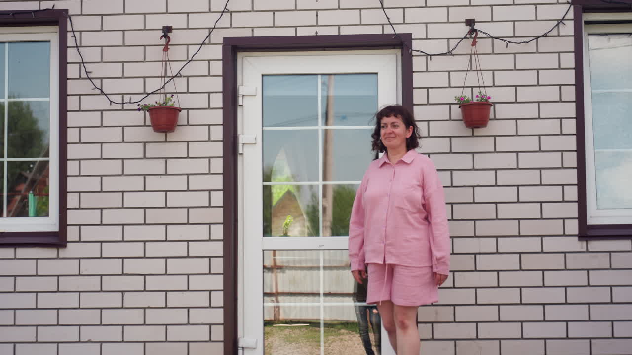 Woman At Door, Pinkshirted Woman Near Brick Entrance, Relaxed Woman Standing By Wall With Hanging Lights, Serene Female Figure In Pink Blouse Beside Floral Planters And String Lighting