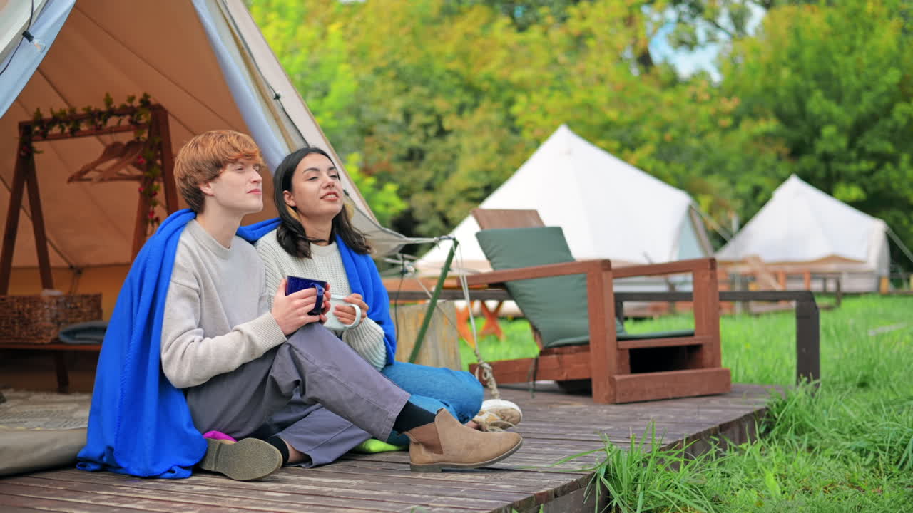 A couple sitting under a blanket on a tent terrace, holding cups and talking at glamping. Lush forest around