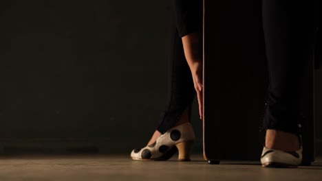 black percussion box in a dark room feet of a woman with black trousers and black and white shoes enter and sit on it