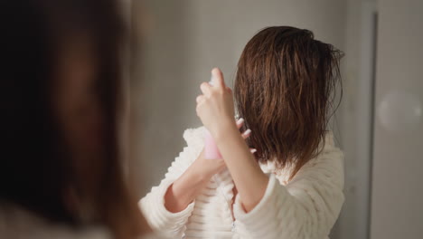 woman with short hair sprays cosmetic product in toilet slow motion. lady in soft terry robe sprays care organic products on hair in light bathroom