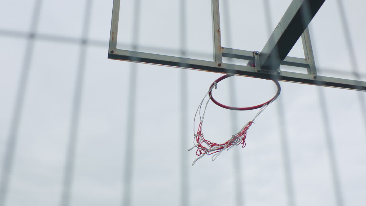 empty outdoor basketball hoop swaying slowly, silent torn net fluttering against overcast sky, chain fence blurred in background, cinematic shallow focus, lonely urban court mood, post practice