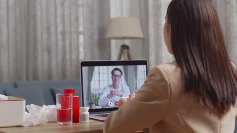 sick asian businesswoman holding a thermometer to the video call with doctor on a laptop while working at home