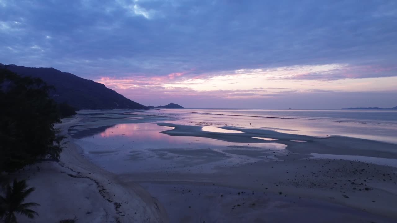 Beautiful aerial footage of sunrise over Koh Phangan in Thailand with calm ocean and pink sky colors reflecting on the beach during low tide