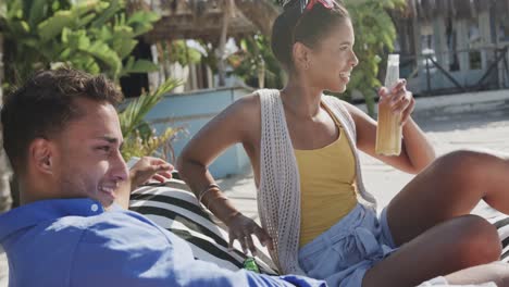 Happy-diverse-couple-talking-and-drinking-beers-relaxing-on-beach-sun-deck,-in-slow-motion