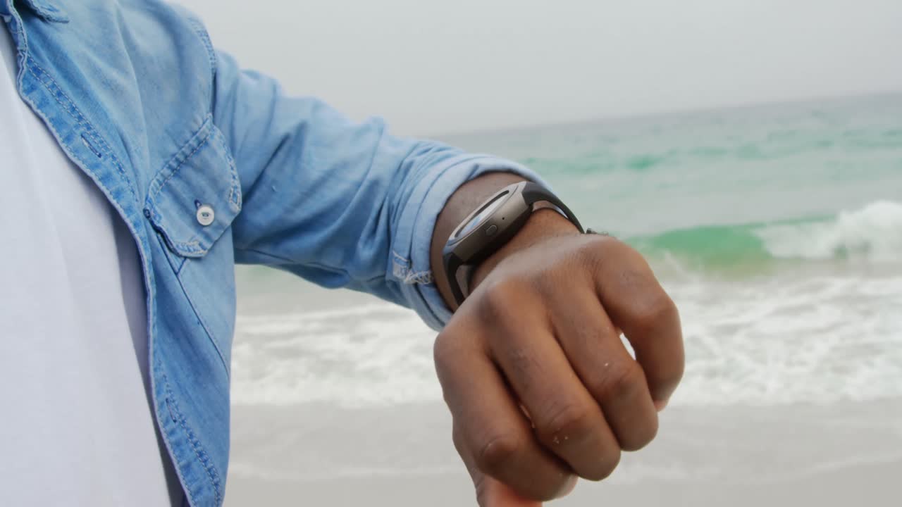 Midtpartiet av afroamerikansk mann som bruker smartklokke på stranden 4K