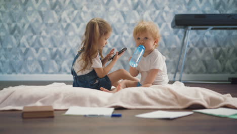 niño con botella se sienta en el suelo cerca de la hermana con teléfono móvil