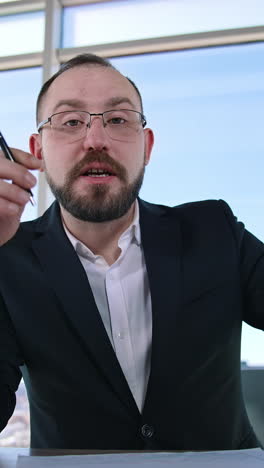 portrait of a businessman sitting in office. bearded entrepreneur in suit and glasses talking about business on window backdrop. vertical video