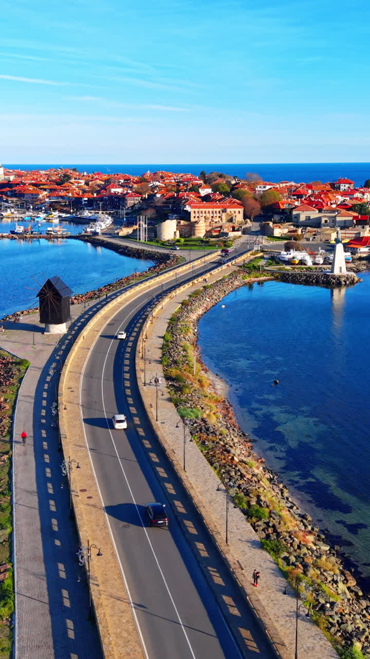 Estrada estreita conectando a cidade velha de Nessebar ao continente, apresentando um moinho de vento tradicional de madeira preta e o Mar Negro. Vista aérea do aterro. Vídeo vertical.