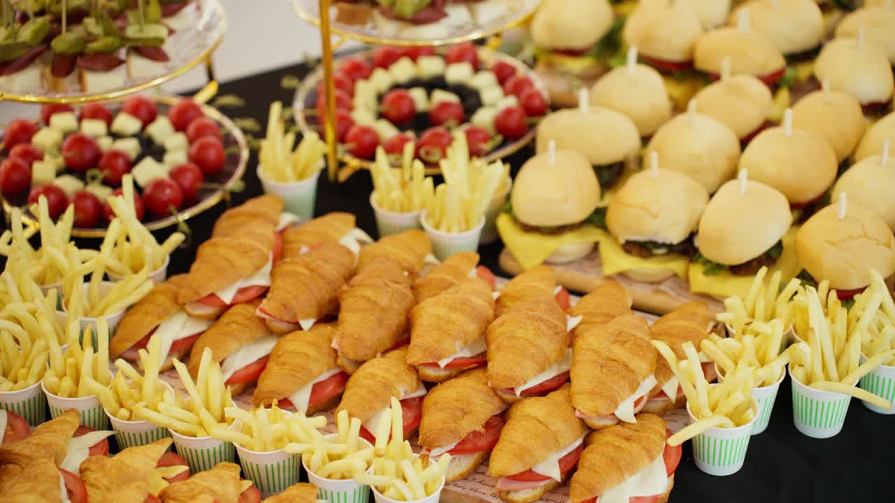 A smooth parallax shot glides across a delicious catering buffet table laden with fresh croissant sandwiches, mini burger sliders, french fries, and other appetizers for a party.