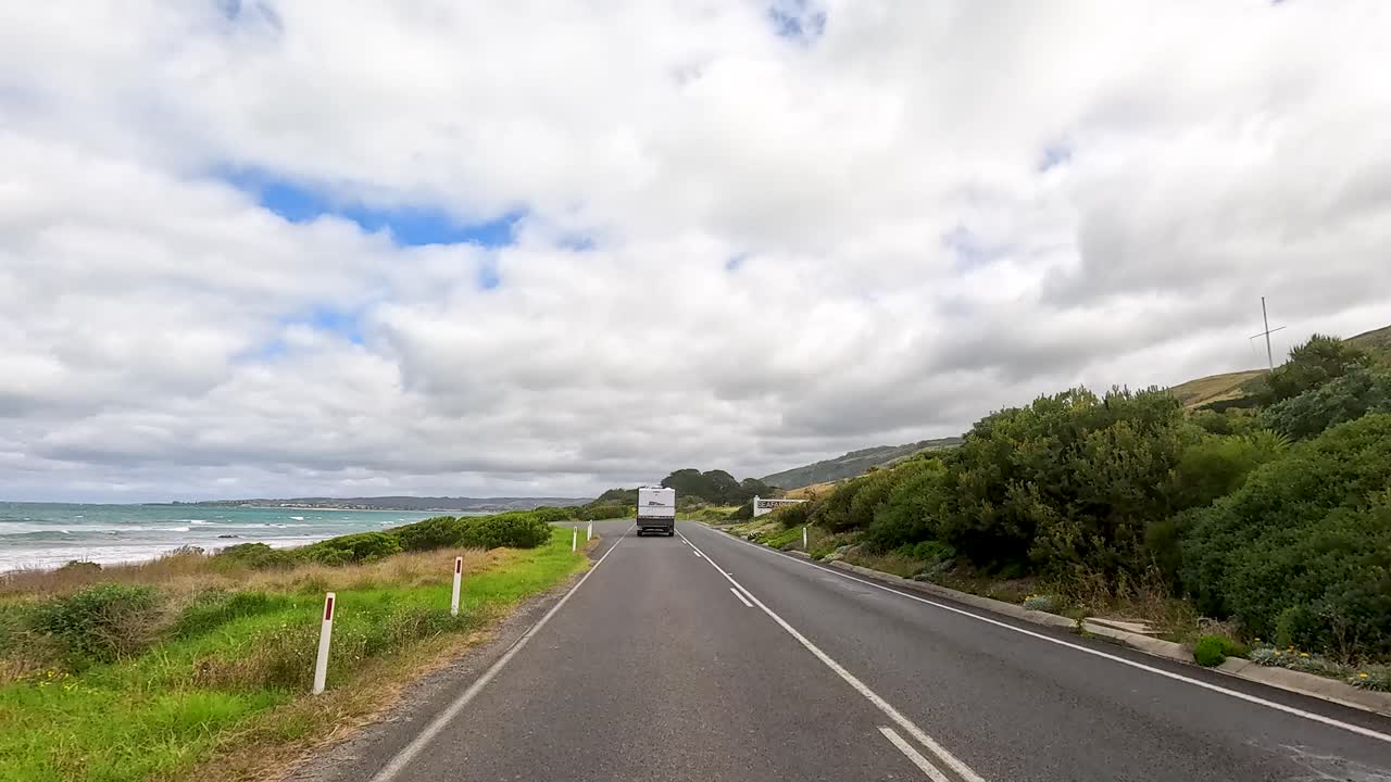A vehicle travels along the picturesque Great Ocean Road, showcasing lush landscapes and coastal views under a partly cloudy sky