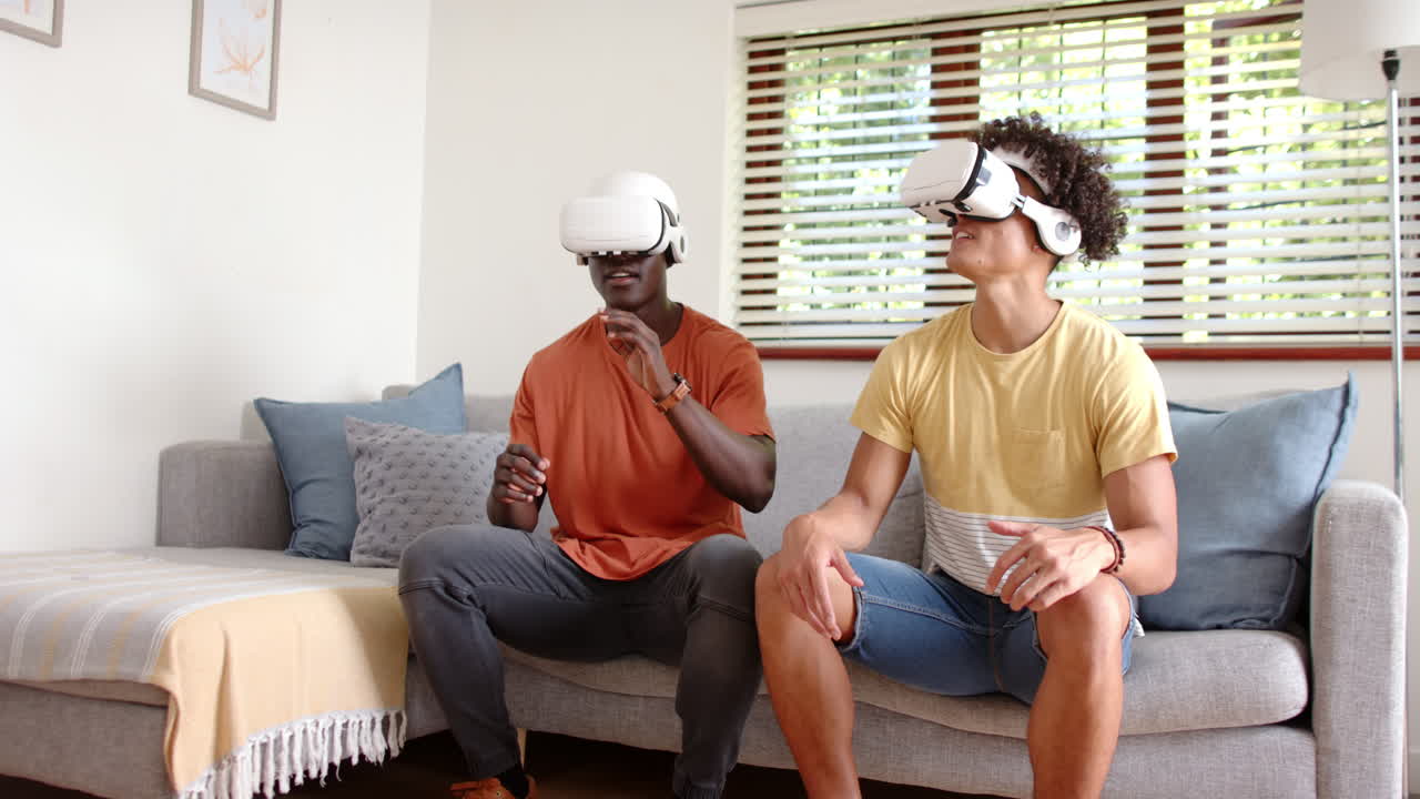 Wearing vr headsets, two multiracial male friends sitting, interacting with virtual world, at home