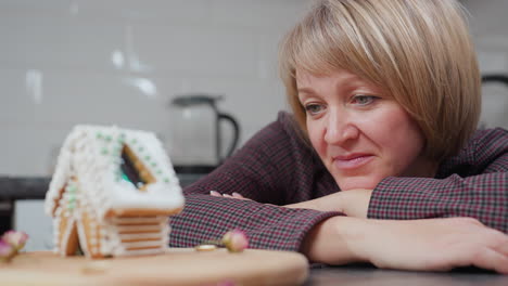 woman rests her head on table, gazing at beautifully decorated gingerbread house with a warm smile, cozy kitchen setting with soft lighting