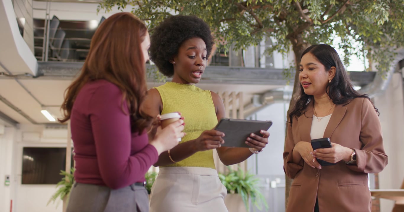 Diverse women in lounge, right speaking on phone, central showing tablet for work, left with coffee