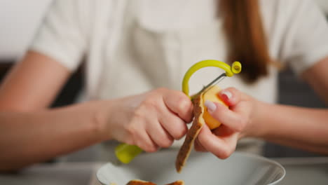 close up of woman using yellow peeler to remove skin from raw potato over bowl in kitchen, hands in sharp focus while background and body remain slightly blurred