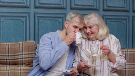 family of senior mother, adult son or grandson relaxing on sofa, enjoying conversation, drinking tea