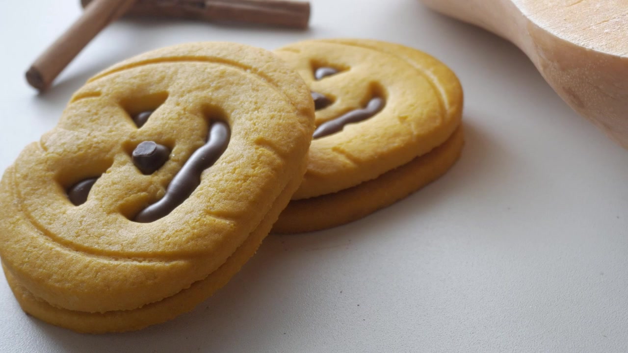 笑顔の顔とチョコレートの装飾が施されたカボチャの形をしたクッキーが白い表面に置かれている。