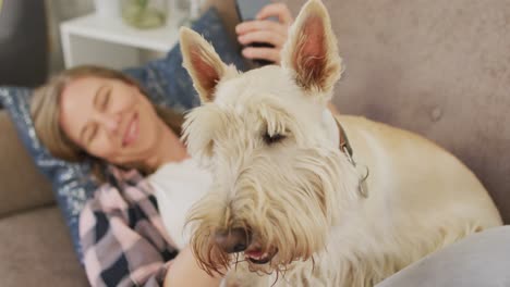 Mujer-Caucásica-Usando-Un-Teléfono-Inteligente-Mientras-Está-Acostada-En-El-Sofá-Con-Su-Perro-En-Casa