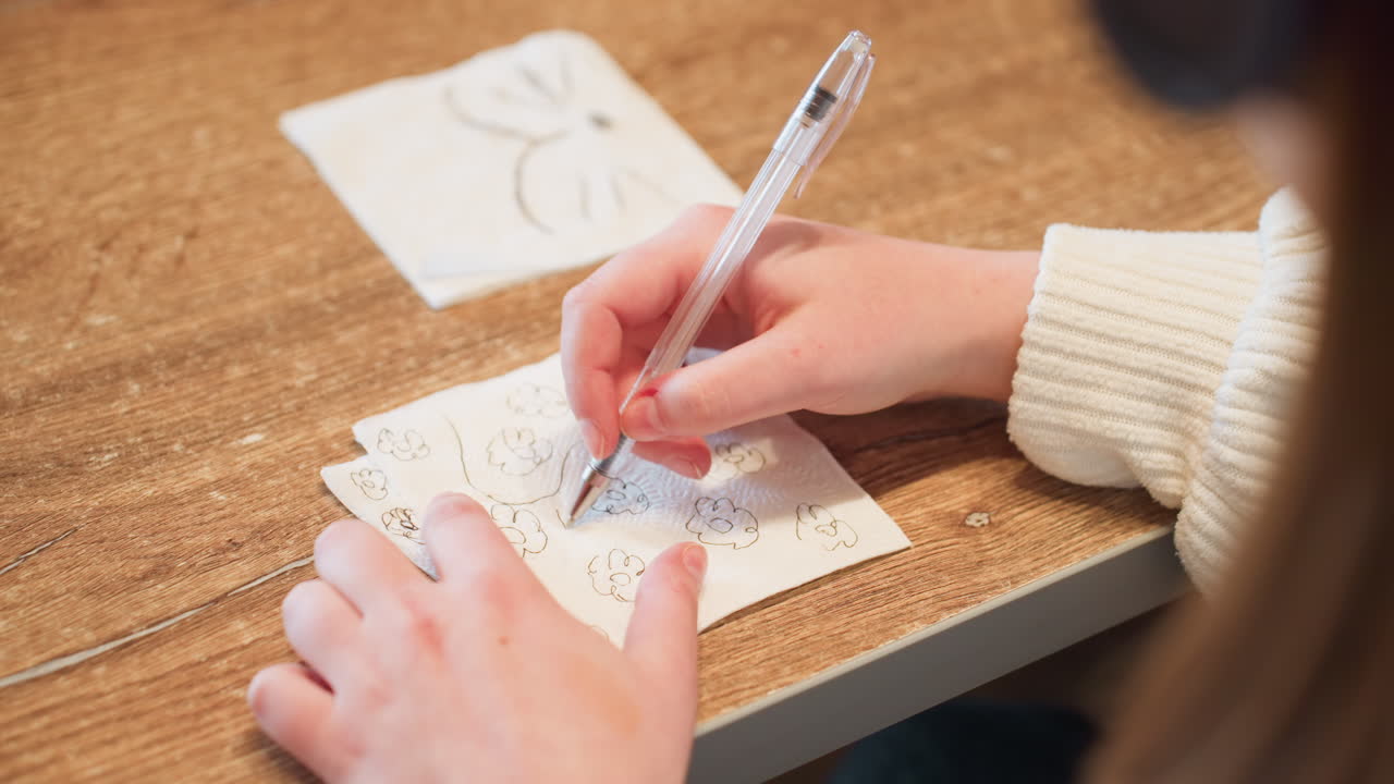 Close up of woman sketching multiple flower designs on tissue paper using pen while seated at wooden table, focusing on hand movements and creative process with warm lighting