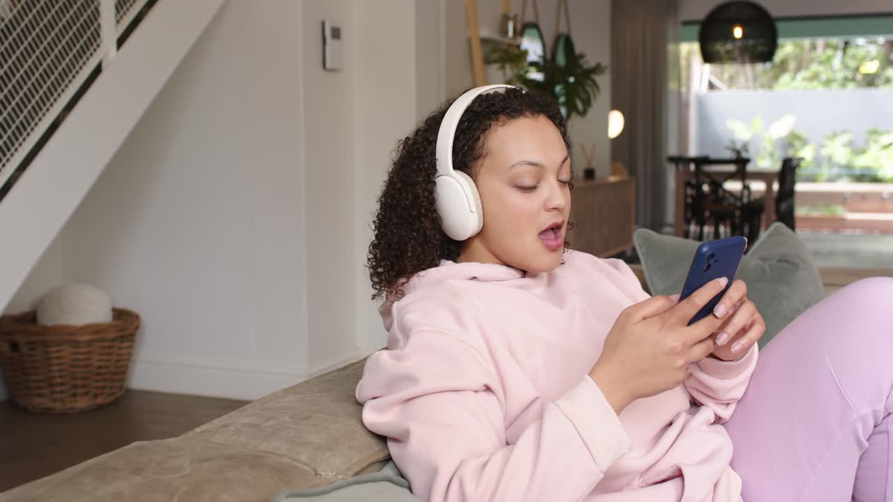 Donna con cuffie bianche che ascolta un audio, scorrendo sul telefono blu sul divano di casa e cantando