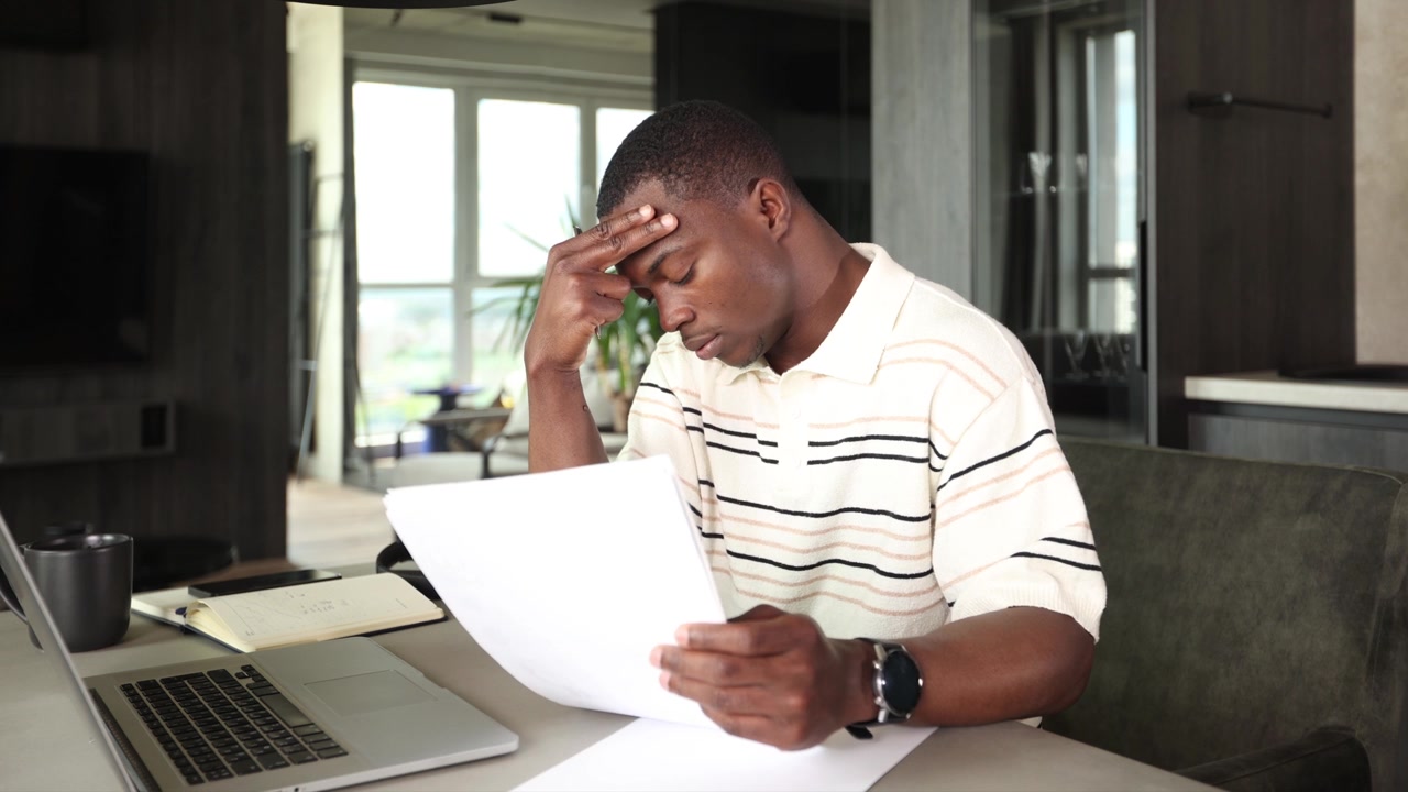 Giovane uomo afroamericano stressato che lavora su laptop e documenti