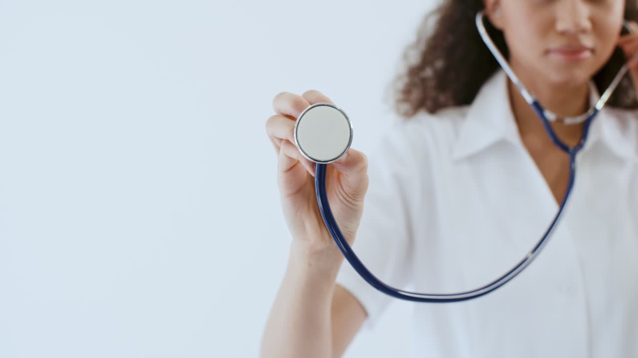 Giovane dottoressa afroamericana in camice medico avvicina lo stetoscopio all'obiettivo della fotocamera in piedi su sfondo bianco in ospedale. Terapista di etnia mista in uniforme. Concetto di assistenza sanitaria e medicina