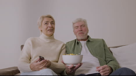 happy senior couple eating caramel popcorn and watching tv while sitting on sofa at home 1