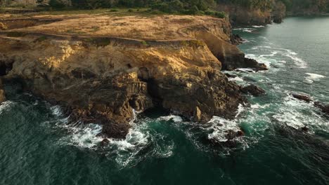 Aerial-view-of-California's-rocky-coastline-at-sunset-with-waves-crashing-into-the-coast