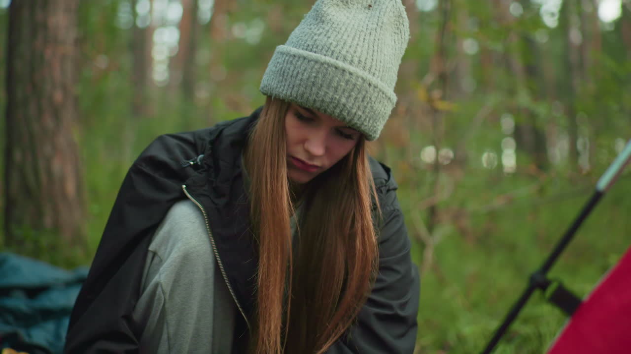lady in black sweater and gray beanie grips wood with both hands hitting ground beside tent in forest setting as trees blur in background and concentration shows on her face