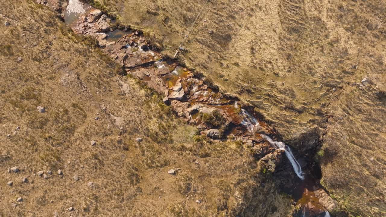 Aerial drone footage of a scenic river flowing through the rugged landscape of the Isle of Skye, featuring a beautiful waterfall surrounded by dramatic hills and untouched nature
